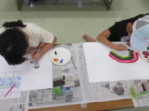 テーブルの上に新聞紙を敷いて白い画用紙に絵具で絵を描いている2人の子どもの写真