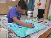 女の子がテーブルの上に新聞紙を敷いてほぼ出来上がっている水彩画に細い筆のようなもので微調整を行っている様子の写真