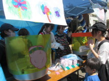 青色のテント内で、関係者たちが親子連れの来場者にわたあめを販売している写真