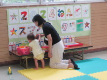 長テーブルにつかまり立ちをした幼児と隣に立つ女性がテーブルで遊べる遊具で遊んでいる写真