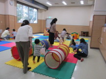 ジョイントマットの上に置かれたキッズテントトンネルの遊具で、楽しそうに遊んでいる幼児や、その様子を見ている保護者の写真