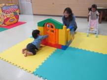 両ひざをついて小さな家のおもちゃのドアを開けようとしている子どもと、その様子を見守る笑顔の女性の写真
