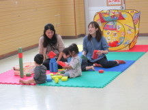 ジョイントマットの上で積み木遊びをしている3名の幼児と、その様子を見ている2名の女性の写真