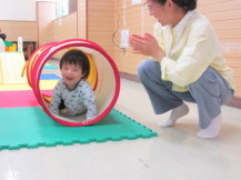 ジョイントマットの上に置かれたキッズテントトンネルの遊具をくぐっている子供と、その横で拍手をしている女性の写真