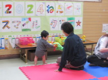 座卓テーブルの上に置かれたおもちゃを手に取る小さな子どもと、その様子を見守る女性の写真