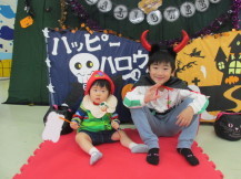 ハッピーハロウィンと書かれた工作のフォトスペースの前で、笑顔で記念撮影している様子の写真