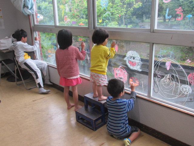 青と水色の縞模様を着た男の子と黄色い服を着た女の子とピンクの服を着た女の子が並んで水で流せるクレヨンで窓にキャラクターや花を書いている写真