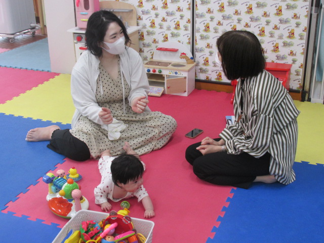 マットが敷かれた部屋で乳児の子育てについて相談する母親とそれについて応答をする相談員の写真