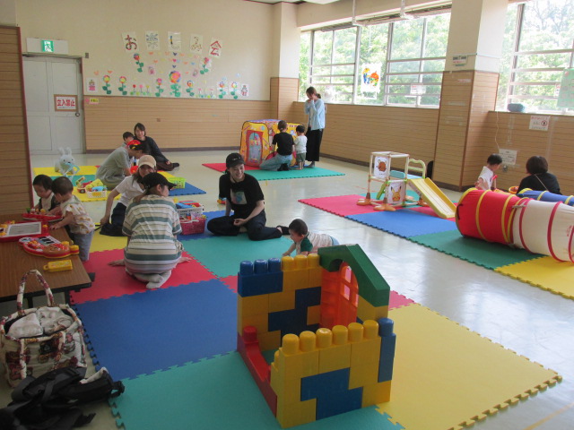複数の親子がマットが敷かれた部屋で乳幼児とともにピアノの玩具やボールプール、人形の玩具で遊んでいる写真