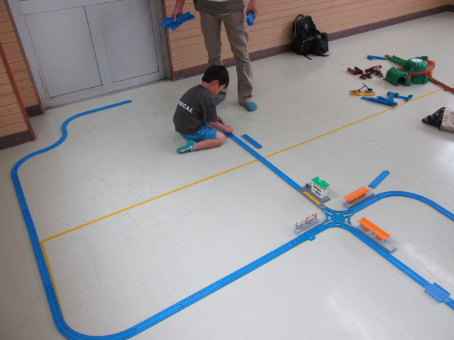 灰色の半そでシャツを着てレールの接続をする男の子とその様子を立って見守る父親の写真