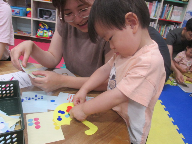 お母さんの膝に乗りながらシールを貼っていく幼児と横で座って作業する子供の写真