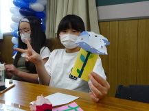 椅子に座った女の子が出来上がった黄色に白色の花のメッセージカードをこちらに向けてピースをしている写真