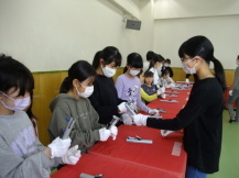黄緑色の床の室内で、横一列に並べられて赤い布が敷かれた長机の前に白い手袋をして並んでいる子どもたちと、その前で黒色の服を着た人がベルの扱いを教えている写真
