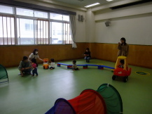 黄緑色の床の室内で、保護者の方に見守られながら車の玩具や青色の平均台のような遊具で遊んでいる子どもたちの写真