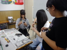 新聞紙の敷かれたテーブルの周りで、3人の女の子が鉛筆を手動で削っている写真