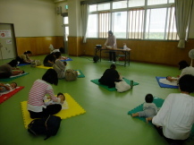 緑色の床にカラーマットの敷かれた室内で、前方に立っている講師の方を見ながら赤ちゃんにベビーマッサージをしている保護者の方々の写真