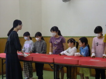 黄緑色の床の室内で、横一列に並べられて赤い布が敷かれた長机の前に白い手袋をして並んでいる子どもたちと、その前で紺色の服を着た人が話をしている写真