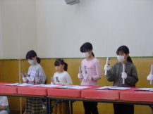 横一列に並んだ赤い布が敷かれた長机の前に、白い手袋をして並んでいる子どもたちが目の前に置かれた楽譜を見ながら練習をしている写真