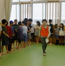 緑色の床の室内で、メモを手に持ち参加者のたくさんの子どもたちを整列させている、オレンジ色のビブスを着た子どもの写真