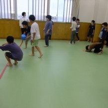 緑色の床の室内で、青色のボールを使ってドッジボールをしている子どもたちや、周りで観戦している子どもたちの写真