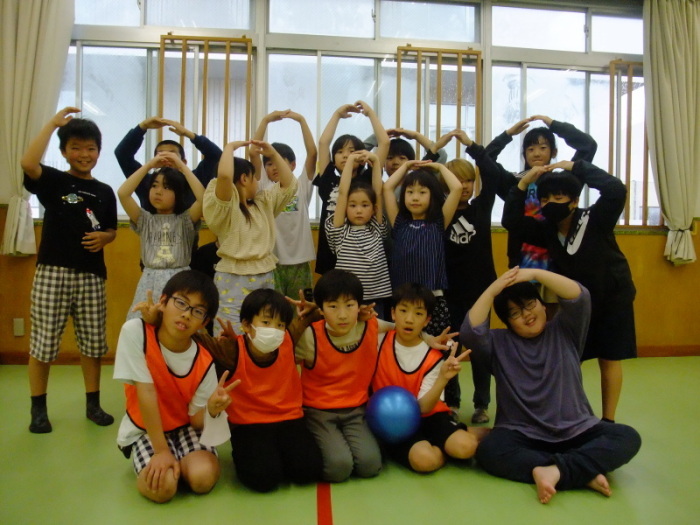 薄緑色の床の室内で、前のほうでオレンジ色のビブスを着てピースをしている運営の4人の子どもたちと、その後ろで手で丸を描いている子どもたちの集合写真