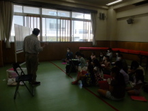 緑色の床の室内で、前のほうに立ちホワイトボードを使用して話をしている講師の先生と、床に座ってそれを聞いている子どもたちの写真