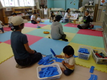 カラーマットの敷かれた部屋で前のほうで絵本の読み聞かせを行っている先生とそれを聞いている子どもたちと保護者や、手前のほうで青いプラレールで遊んでいる赤ちゃんの写真