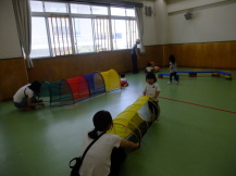緑色の床の室内で、奥のほうで子どもが乗って遊べる車の玩具や、トンネルなどの普段は見慣れない遊具で遊んでいる子どもたちとそれを見守っている保護者の写真