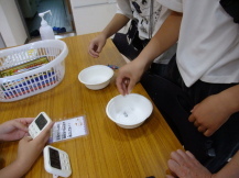 テーブル上で白い器にサイコロを振っている子どもと、それを見守ったりタイマーで時間を図っている子どもたちの手元の写真