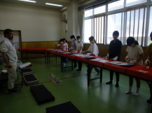 緑色の床の室内で、横一列に並べられた赤い布が敷かれた長机の前で、ハンドベルを手元に持ちながら配布された楽譜を見ている子どもたちの写真