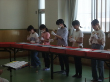 緑色の床の室内で、横一列に並べられた赤い布が敷かれた長机の前で、配布された楽譜を見ながらハンドベルを鳴らして練習をしている子どもたちの写真