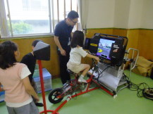 緑色の床の室内で、映像を映すモニターが正面に設置された自転車シミュレーターで担当の方が隣につきながら実際に自転車部分をこいでいる女の子と、それを見ているほかの子どもたちの写真