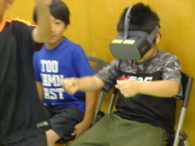 VR体験用の大きな機械式の眼鏡のようなものを付けた子どもが椅子に座りながらハンドルを握っているような動作をしている様子の写真