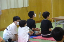 ゲームをしている子供の後ろでこそこそ話している2人の男の子の写真