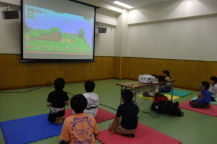 夢中になってゲームを遊ぶ4人の子供たちと後ろで観戦する子供たちの写真