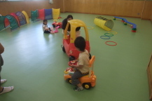 広い室内で車の遊具に乗って遊んでいる子供たちを撮影している写真