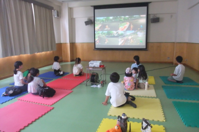 スクリーンに映し出されるゲームを座って楽しむ子供たちの写真