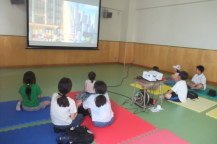 ゲームを楽しむ子供と後ろに座って観戦する子供たちの写真