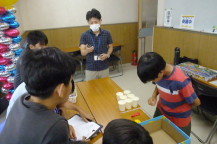 長テーブルを組み合わせて2列に並べその周りを子どもたちや関係者が囲み1人の子どもがテーブルに置かれた紙コップの前で左手を後ろに回して前かがみになっている写真