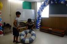 風船で装飾された室内で2名の子どもが立っており、そのうちの1名が浮き輪を持とうとしている写真