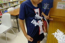 長テーブルの上に置かれた沢山の洗濯ばさみの前で子どもが自分が着ているTシャツにも沢山の洗濯ばさみをつけている写真