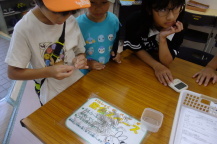 テーブルの前に立っている3名の子どものうち1人がクリップを両手でつなぎ合わせようとしておりその様子を残りの2名が見守っている写真