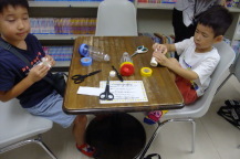 はさみやペン、ペットボトルなどが置かれたテーブルの前に対面に座った2名の子どもが制作を行っている写真