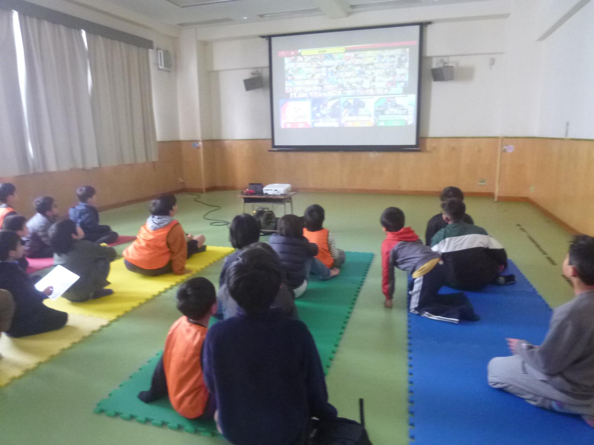 集会室のスライドにゲーム画面を映し、遊ぶ様子。20人ほどの子どもたちが順番を代わりながら遊ぶ。