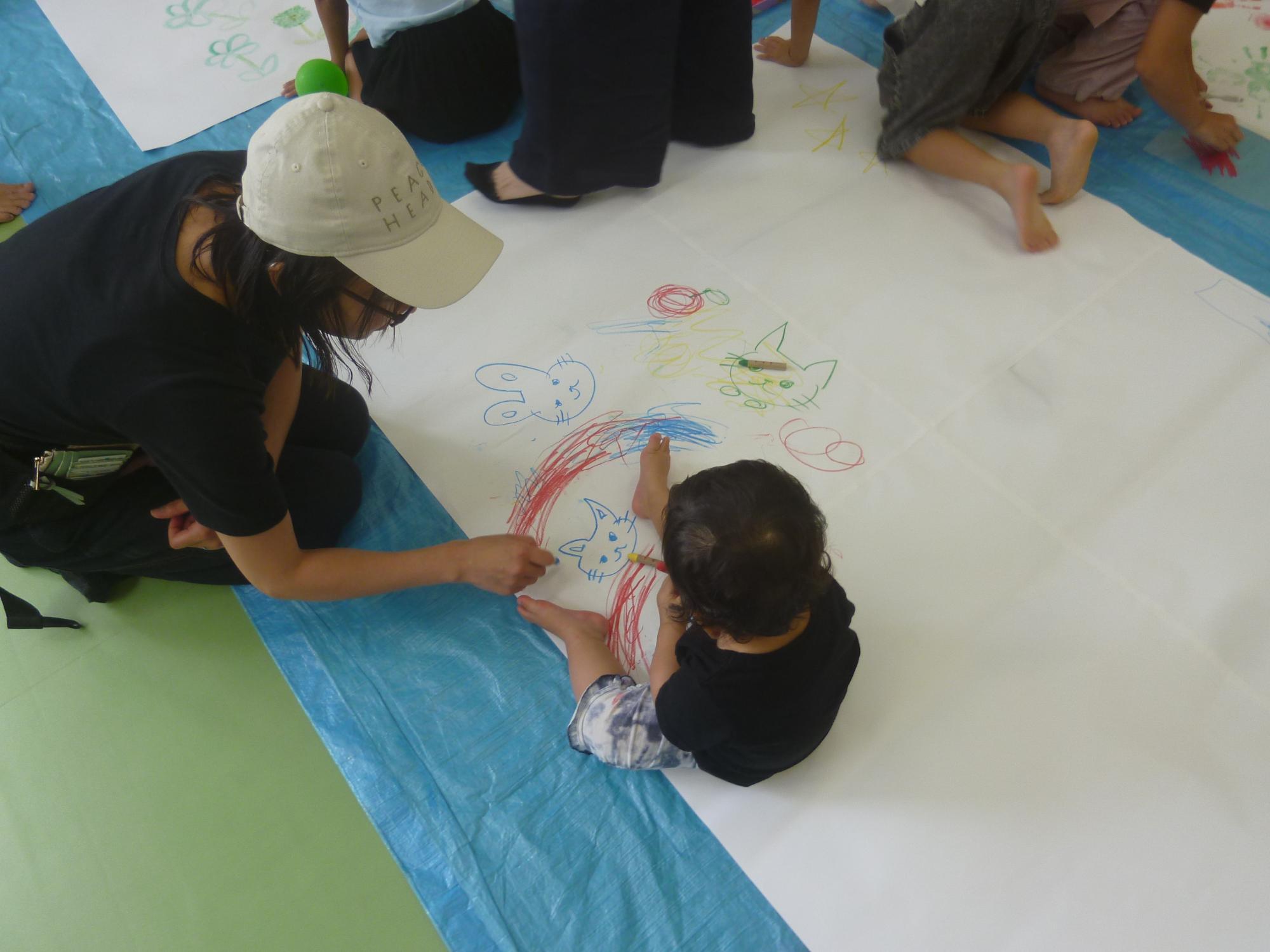 「お絵描き広場」母親とともに大きな虹の絵を描く幼児の写真