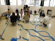 集会室の床に広げて置いたプラレールを、沢山の親子連れが眺めている様子の写真