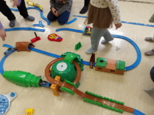 子供たちが座ったり歩いたりしている床に設置された、完成したプラレールをアップで撮影した写真