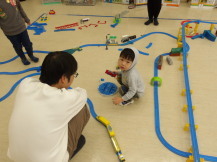 保護者に見守られながら電車で遊んでいるグレー色のパーカーを着た男の子の写真