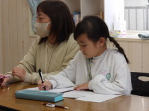 女性職員の隣に座る書記担当の女子児童が、こども運営会議で出た意見をまとめている様子の写真