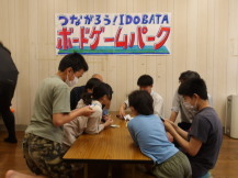 「つながろうIDOBATAボードゲームパーク」と書かれた横幕が壁に貼られた室内で、長机の両側に座ってボードゲームを楽しんでいる小学生や大人たちの写真